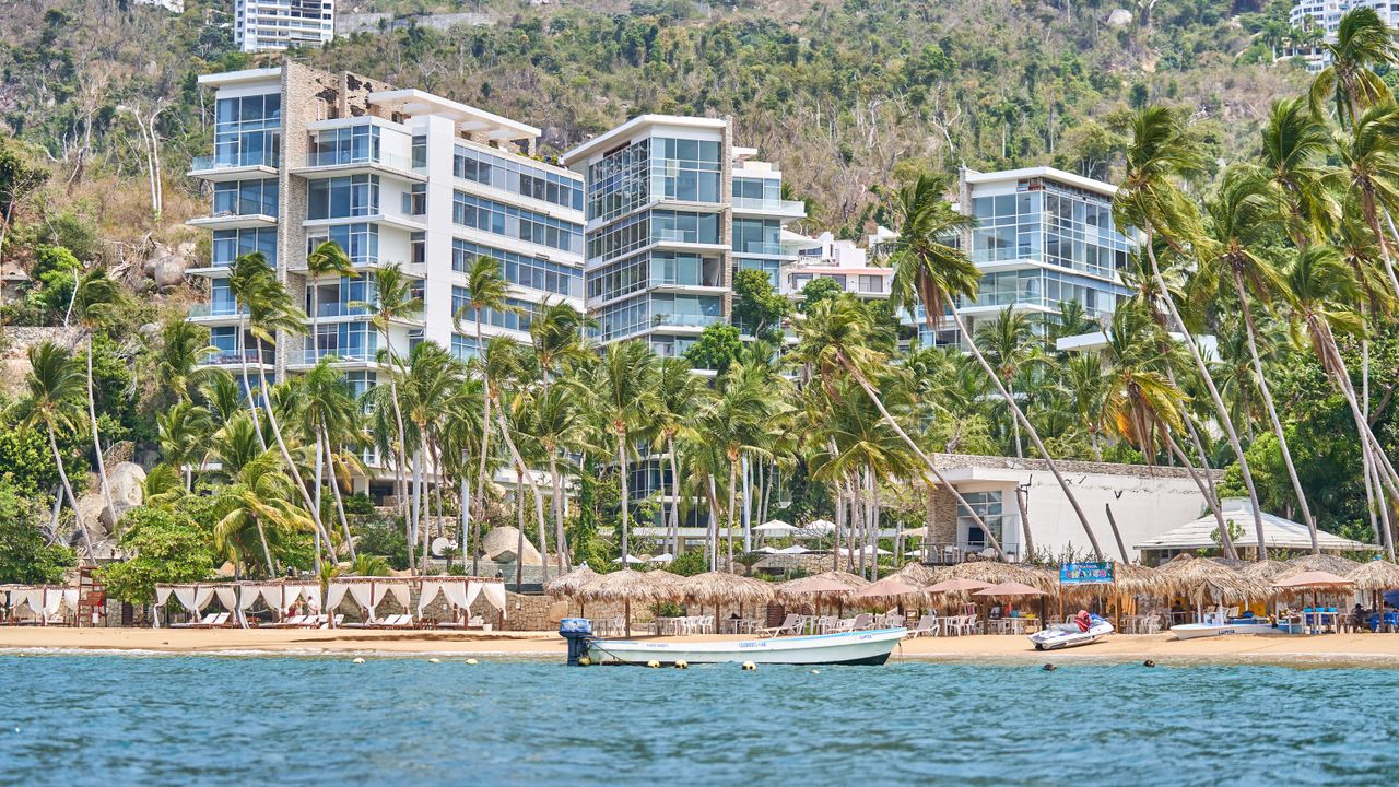 our-story-02 Modern beachfront apartments amidst palm trees along a tropical shore.